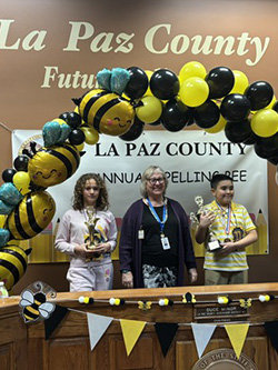 Two students with their spelling bee trophies posing with an adult in front of the La Paz County sign